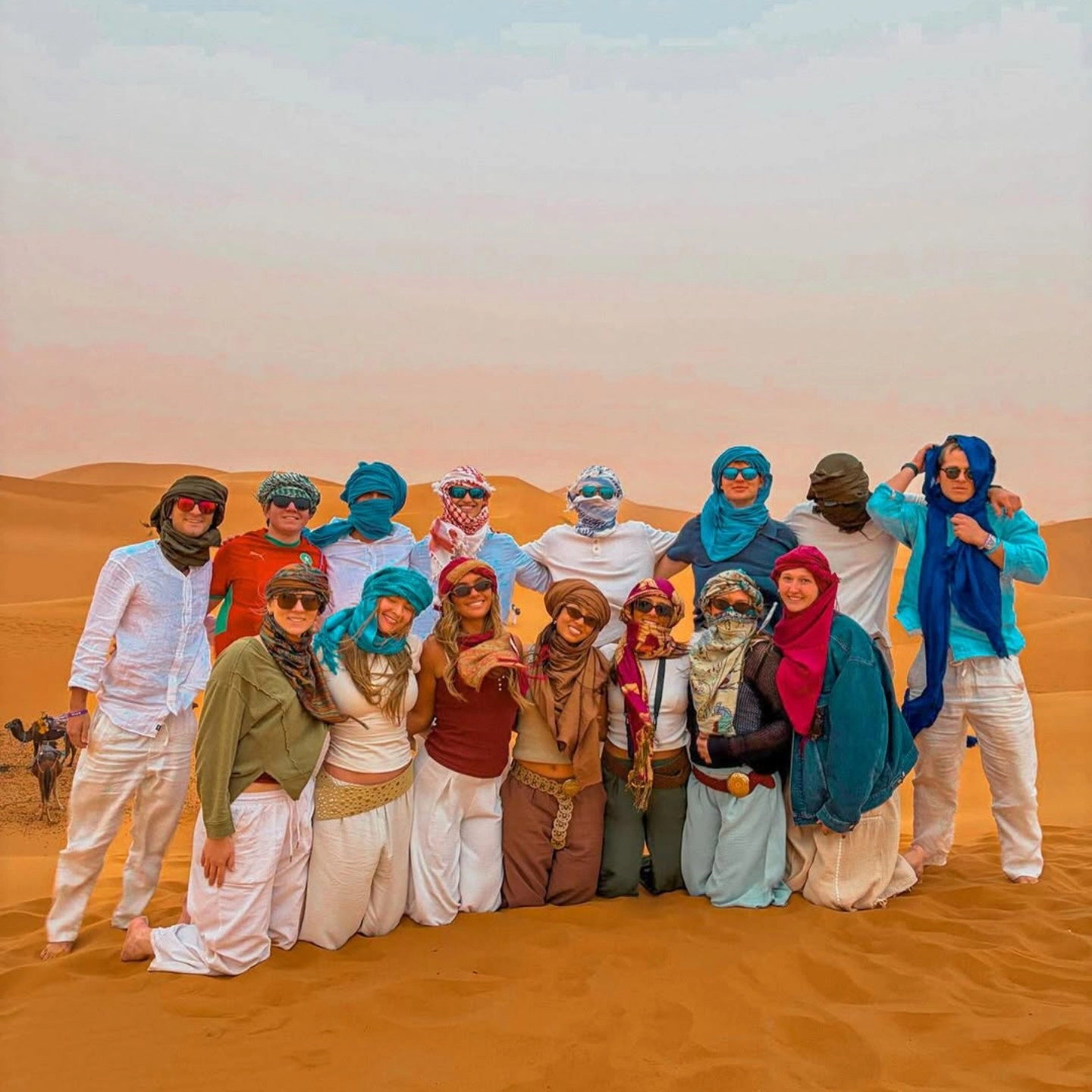 3 days desert tour from Marrakech — large group of travelers wearing colorful Berber turbans on Erg Chebbi dunes Merzouga Sahara Desert Morocco at sunset