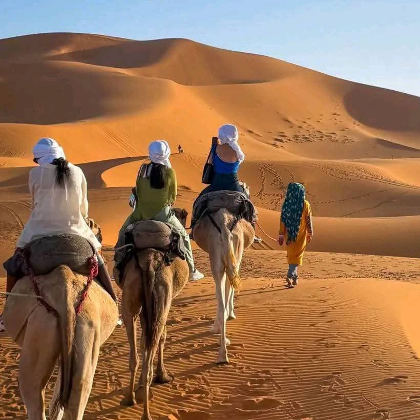 4 days desert tour from Marrakech — group of three travelers riding camels into Erg Chebbi dunes led by Berber guide in yellow robe Merzouga Sahara Desert Morocco