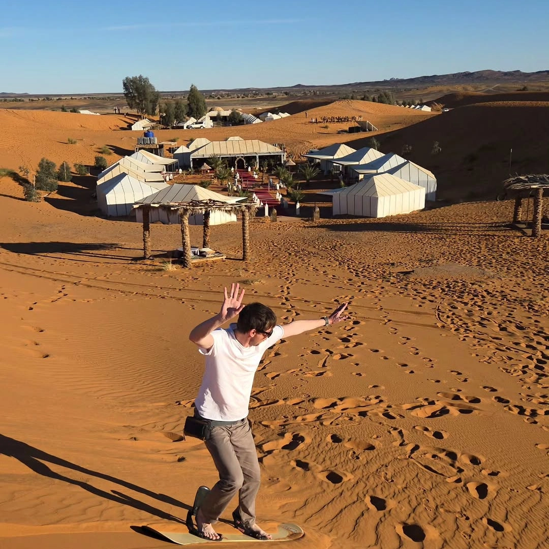 4 days desert tour from Marrakech — man sandboarding down Erg Chebbi dune with luxury Berber desert camp visible below Merzouga Morocco