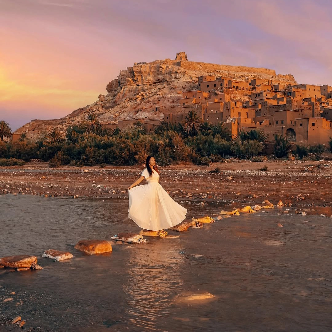 3 days desert tour from Marrakech — Aït Benhaddou UNESCO World Heritage kasbah at golden sunset woman crossing river stepping stones Day 1 Morocco