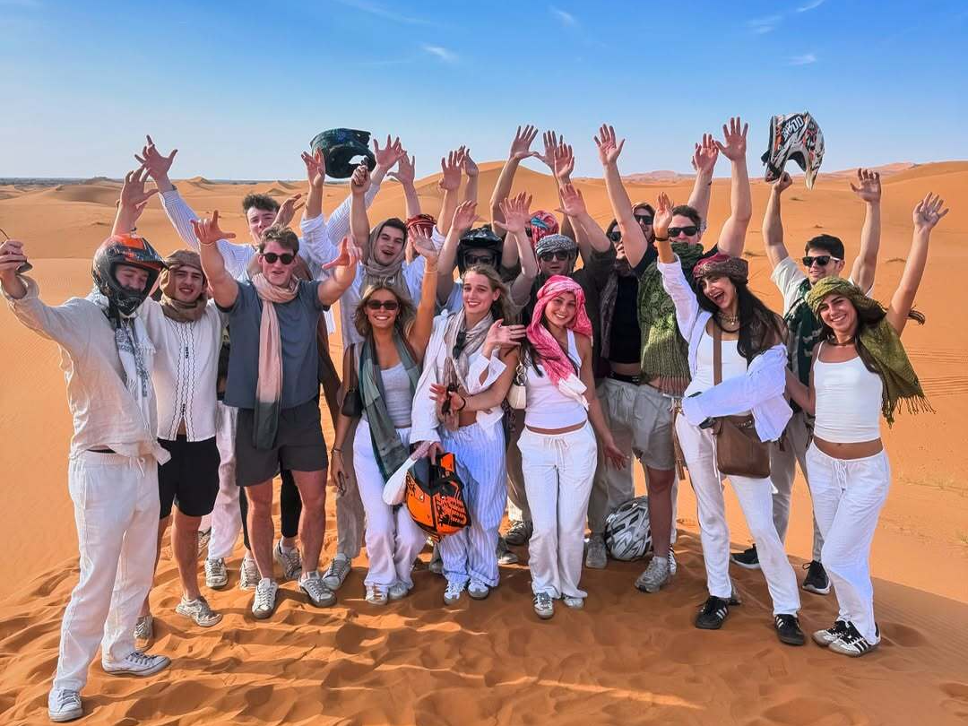 Marrakech to Fes desert tour 3 days — large group of travelers celebrating with arms raised on Erg Chebbi dunes after quad biking Merzouga Sahara Desert Morocco