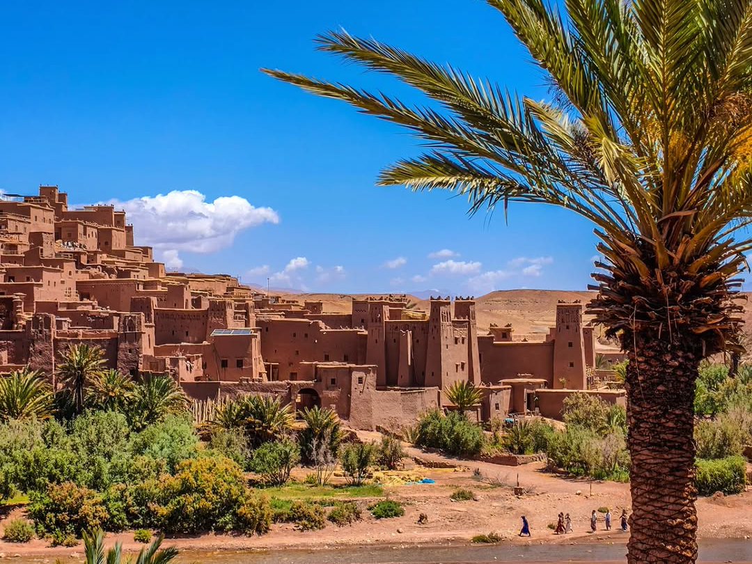 Panoramic view of the ancient mud-brick village of Ait Benhaddou across the Ounila River with date palms in the foreground, Morocco