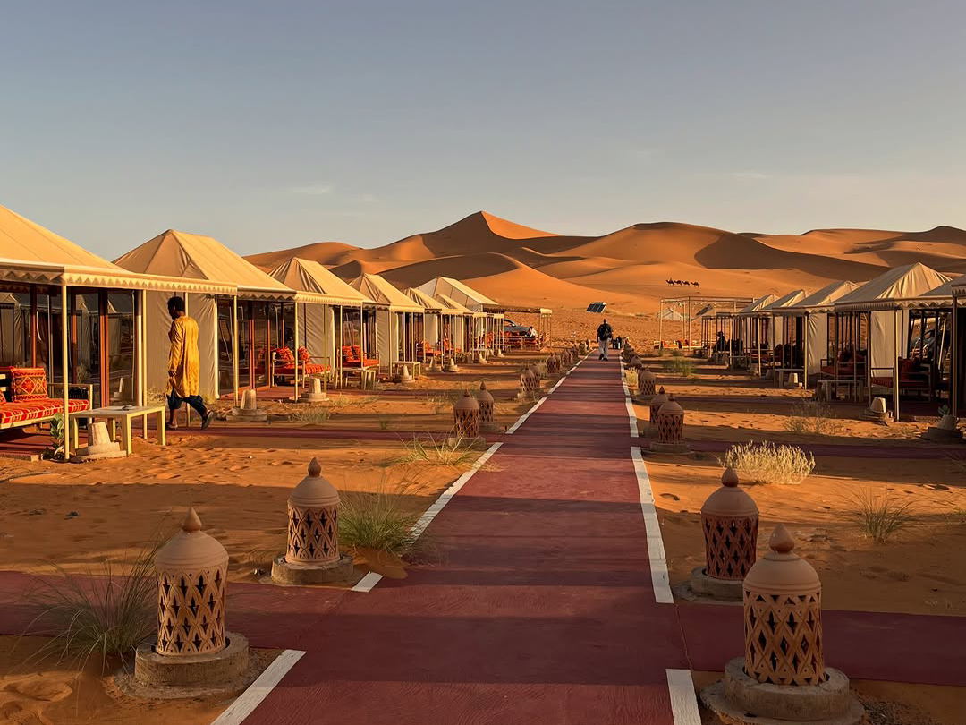 Luxury glamping tents lining a lantern-lit pathway at a Sahara desert camp in Erg Chebbi at sunset, included on the Marrakech to Fes 3-day desert tour