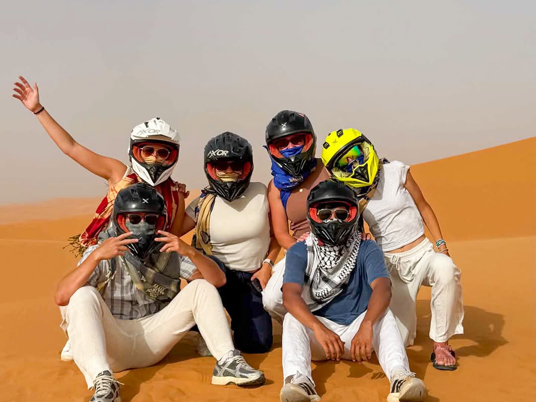 Group of six tourists wearing helmets and face masks posing together on the orange sand dunes of the Sahara Desert after quad biking near Merzouga, Morocco