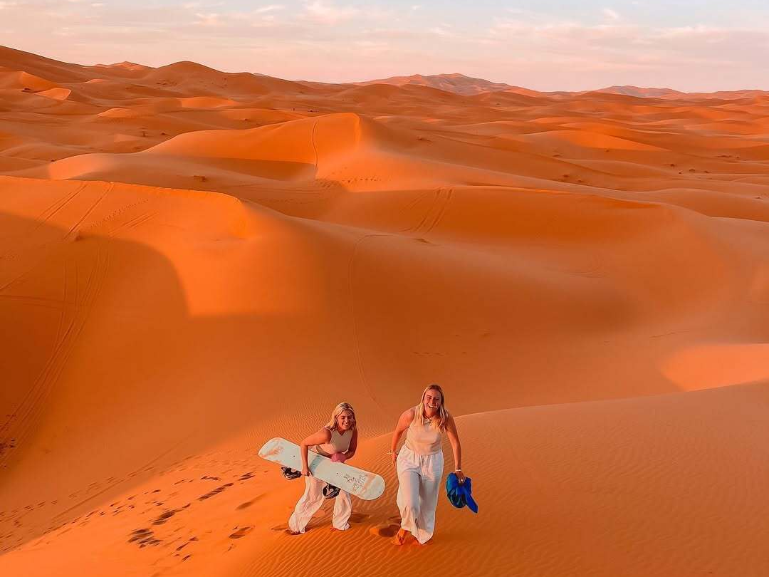 Two women holding a sandboard and walking up the glowing orange dunes of Erg Chebbi at sunset during a sandboarding activity on the Marrakech to Fes desert tour, Morocco